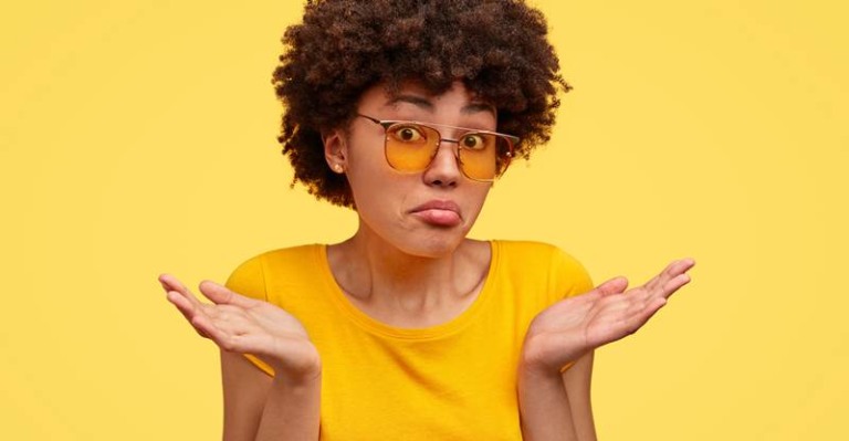 Uncertain dark skinned female shruggs shoulders with puzzlement, dressed in casual yellow t shirt, feels uncertain, makes decision, isolated over yellow background. Woman doesnt care, has no idea