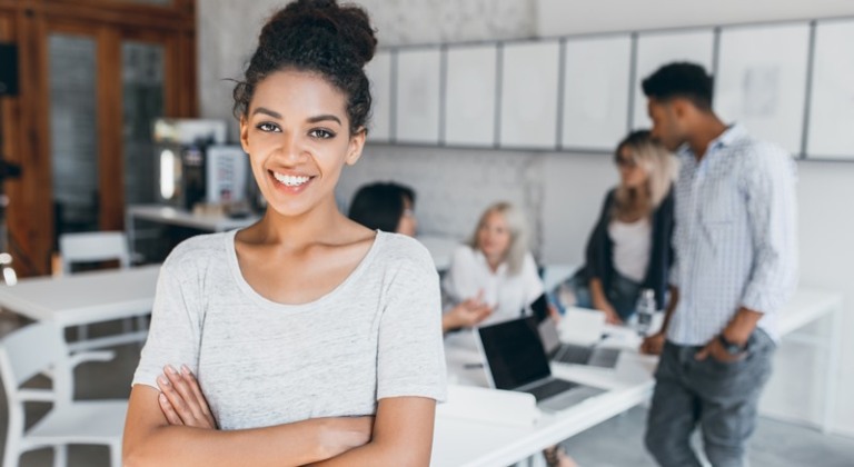 Pleased woman with light-brown skin posing with crossed arms and smiling, while people behind her working. Indoor portrait of tired students with laptop and african curly girl on foreground.