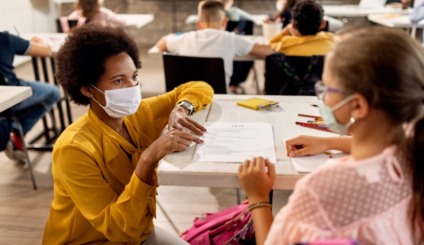 African American teacher and schoolgirl wearing protective face masks while discussing about test results on a class at the school.