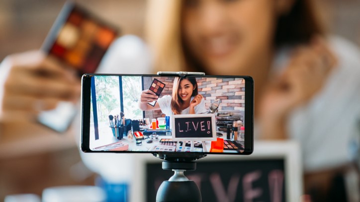 Happy young attractive Asian ethnic model filming vlog on mobile phone on tripod and showing eyeshadows at table with cosmetics