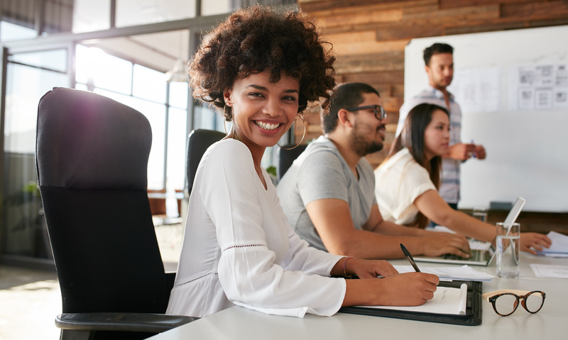 Portrait of happy young african woman sitting at a business presentation with colleagues in boardroom. Female designer with coworkers in conference room.