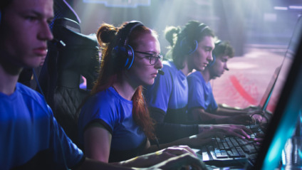 Concentrated young female esportsman gamer in glasses playing shooter on computer while sitting amidst teammates during gaming tournament