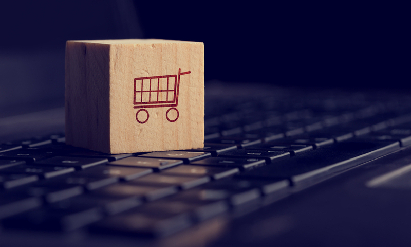 Online shopping and e-commerce background with a wooden cube showing a shopping cart icon resting on a computer keyboard viewed low angle over black with copyspace.