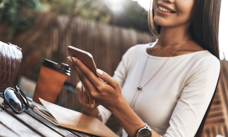 Quick message to client. Close-up of attractive young woman holding a smart phone and smiling while sitting in restaurant outdoors