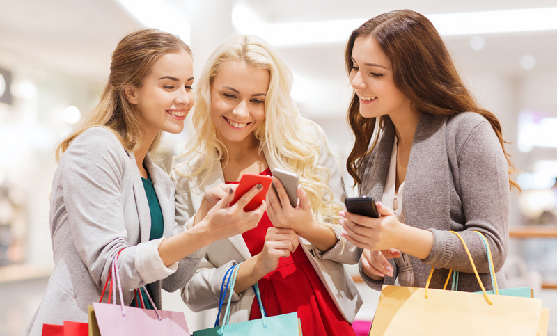 sale, consumerism, technology and people concept – happy young women with smartphones and shopping bags in mall