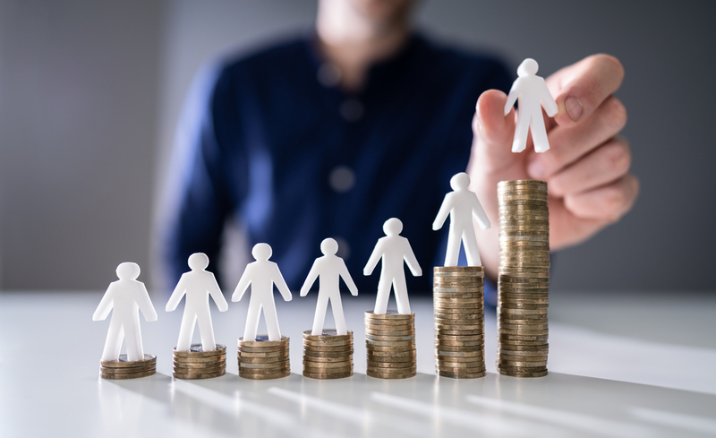 Human Hand Placing Small Human Figure On Increasing Stacked Coins Over Wooden Desk