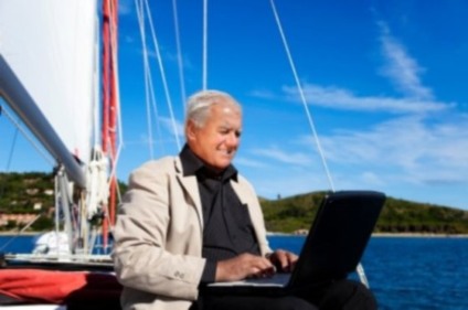 The businessman is working during the vacation on a sailboat with a lovely beach in the background.
