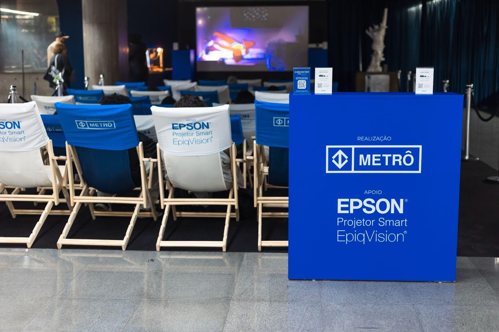 Cadeiras brancas e azuis com o logo da Epson em frente a projeção de um filme oferecido pelo projeto Cinemetrô