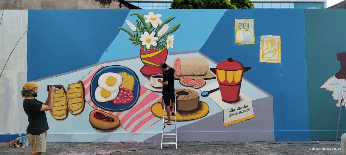 Mural na r.u.a dos amores, em Recife, mostra uma mesa de café posta com bolo de rolo, xícara de café, bolo coberto por castanha de caju, ovos fritos, mamão aberto entre outros alimentos. O artista Rômulo Jackson assina a obra.