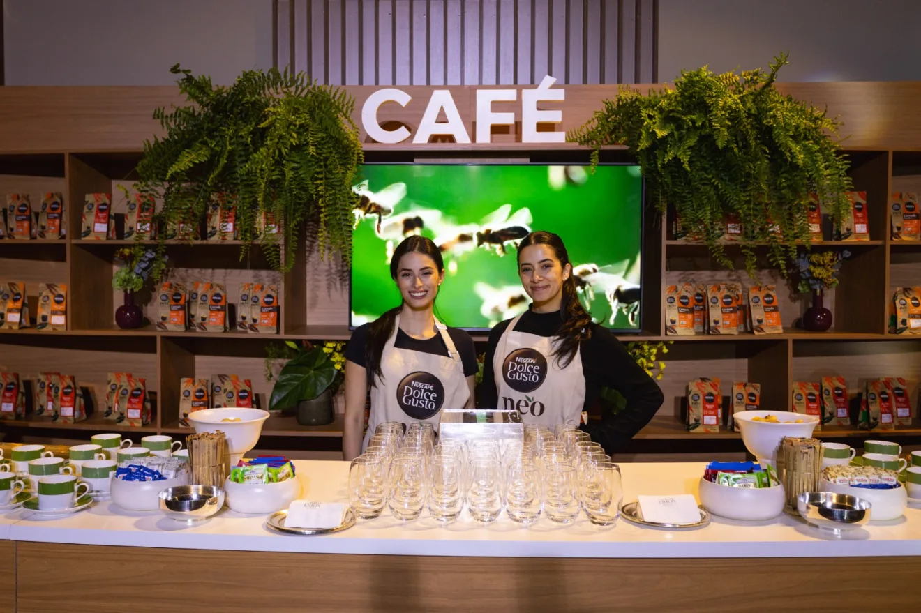 Imagem mostra duas promotoras da marca Dolce Gusto, da Nestlé,no espaço de café do evento. Há uma mesa com xícaras e ao fundo uma estante com folhagens verdes e produtos da marca