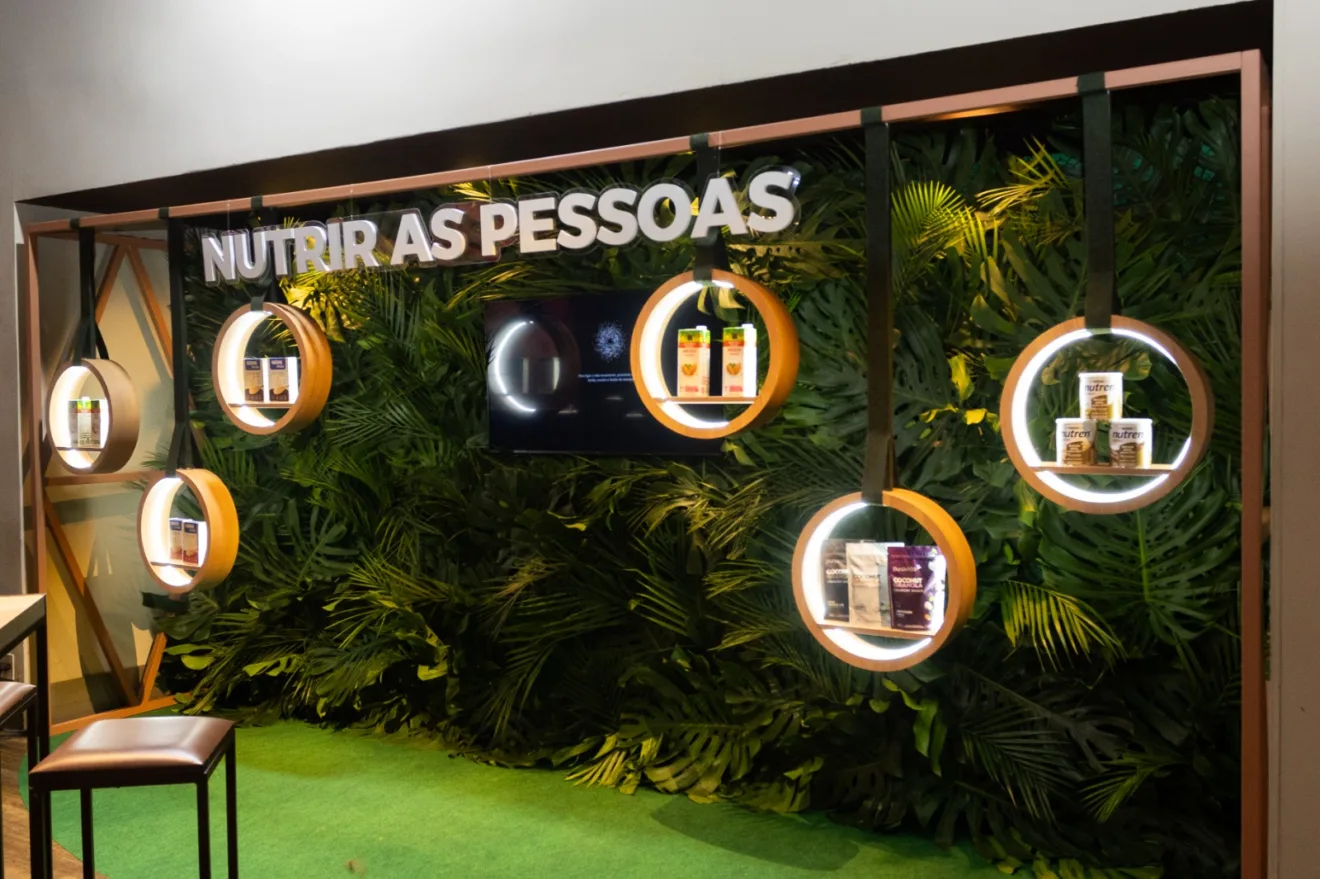 Parede decorativa do evento possui folhagens verdes, com estantes circulares que exibem produtos da Nestlé, e um letreiro no qual está escrito "nutrir as pessoas"