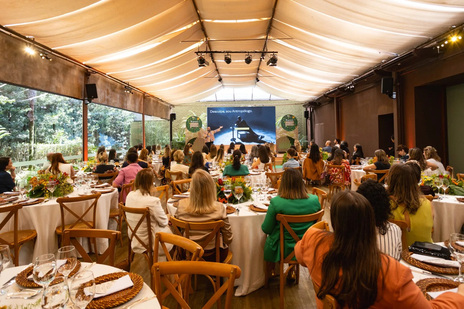 A imagem mostra o espaço do evento Regenerar da Nestle, com convidados sentados em mesas redondas de jantar, em uma decoração amarelo queimado e bege