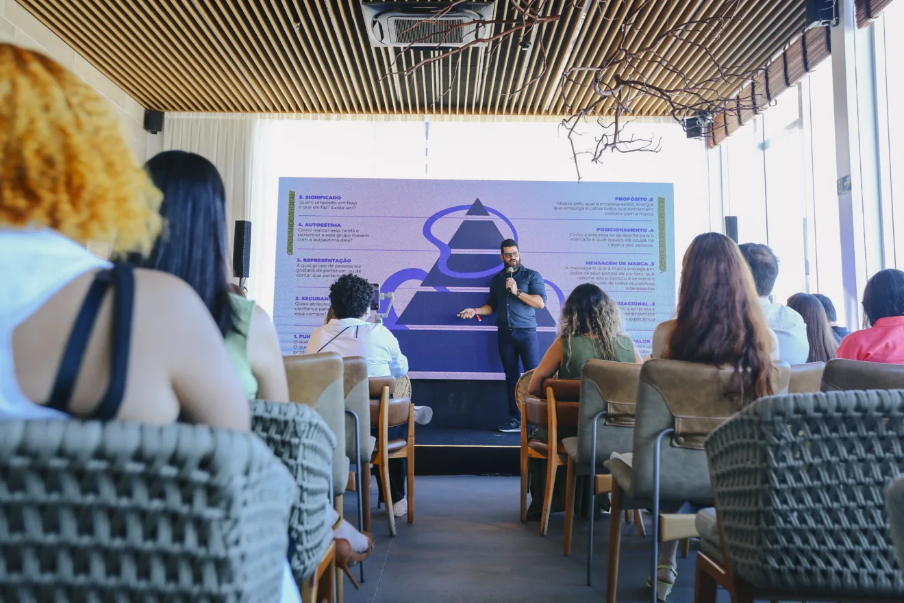 Ricardo Rocha, CEO da agência Energia das Marcas, está em pé, segurando um microfone, palestrando sobre branding. No recorte da foto é possível ver cadeiras com ao menos seis pessoas acompanhando a palestra.