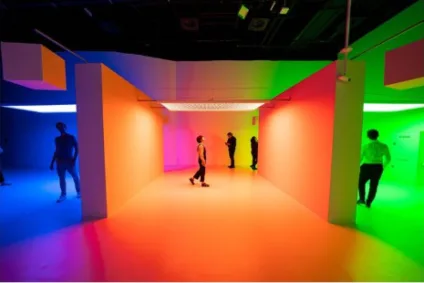 A imagem mostra uma sala de exposição com luzes forte nas cores laranja ao centro, roxa e azul escura à esquerda e verde à direita. O espaço de exposição é como uma sala vazia, com paredes divisórias que não possuem nada além da luz. Há pessoas assistindo a obra.